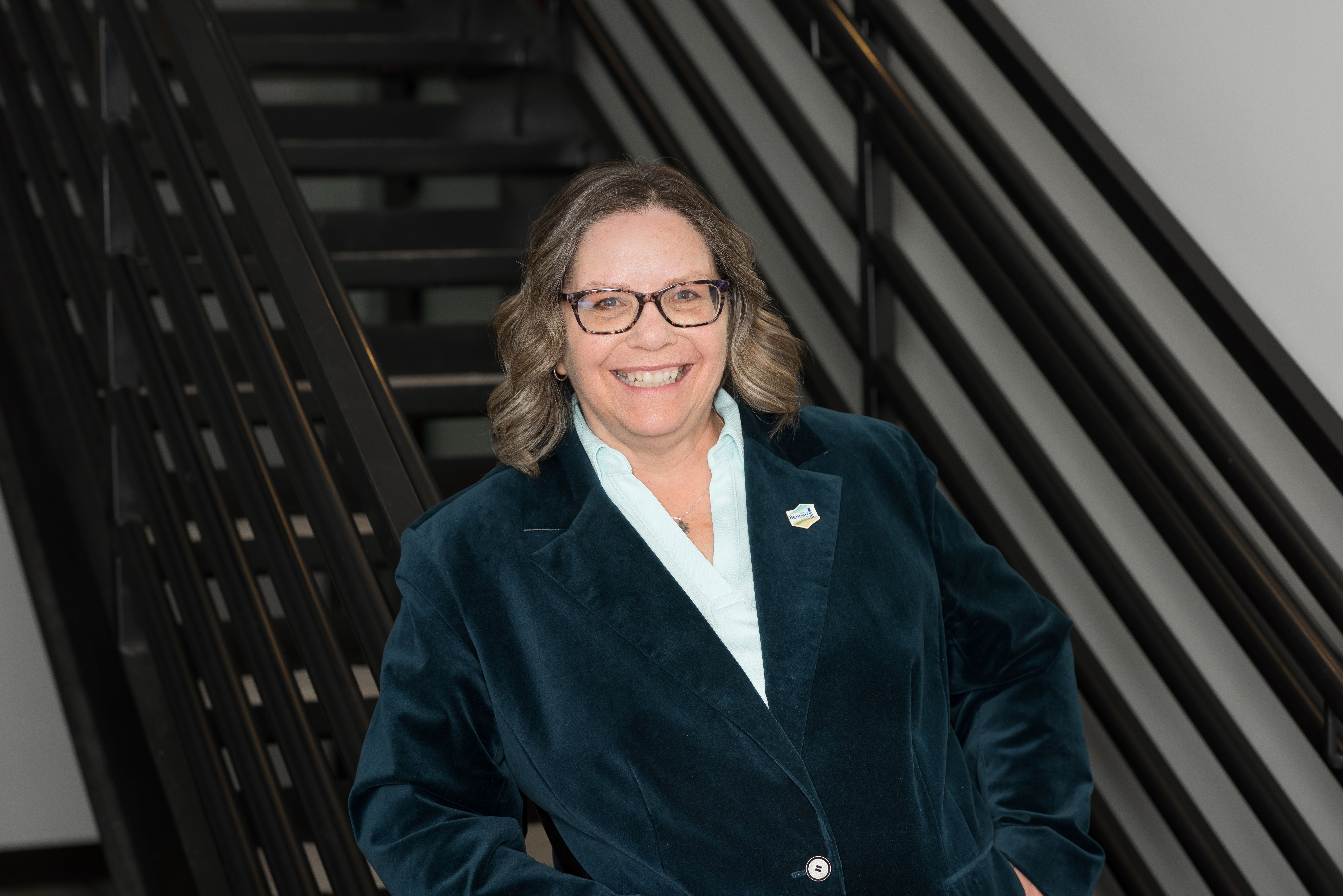 woman with glasses standing by stairs wearing a dark green blazer