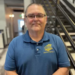 A photo of a while male in classes wearing a blue shirt with a gold town of bennett logo in the corner standing in front of a staircase