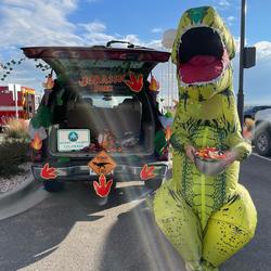 Person in a dinosaur costume standing in front of a decorated trunk at Trunk or Treat event.