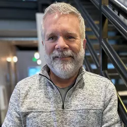 A man with white hair and beard wearing a grey sweater standing in front of black stairs. Greg Thompson planning manager. 