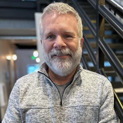A man with a grey beard, smiling and wearing a grey zip up sweater is standing in front of a staircase.