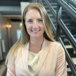 Executive assistant rachel hample. Blonde woman wearing a pink blazer standing in front of stairs