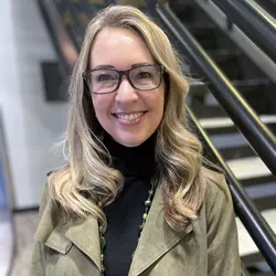 Photo of a blonde woman wearing glasses and a green jacket standing in front of a staircase. Communications Coordinator Tiffany Chaput