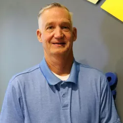 Trustee Steve Dambroski. Man standing in front of blue wall wearing a blue polo shirt. 