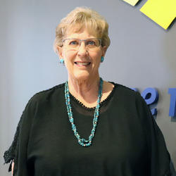 Trustee Donna Sus. Woman standing in front of a blue wall wearing glasses in a black shirt with blue necklace