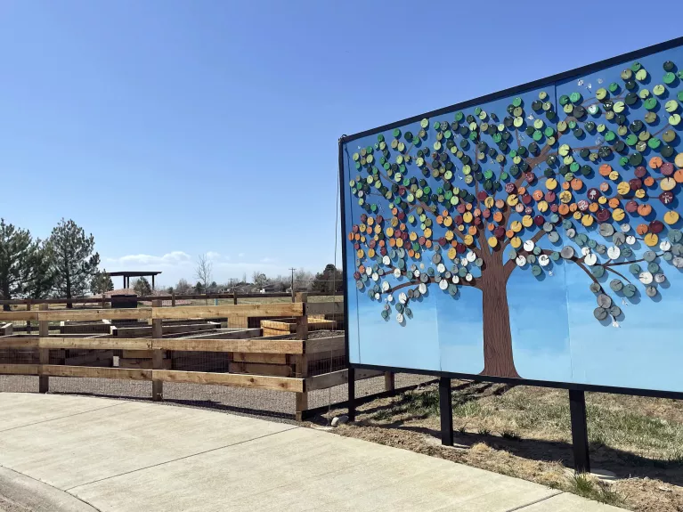 Picture of a mural with a tree and multiple different color leaves and a community garden on a sunny day